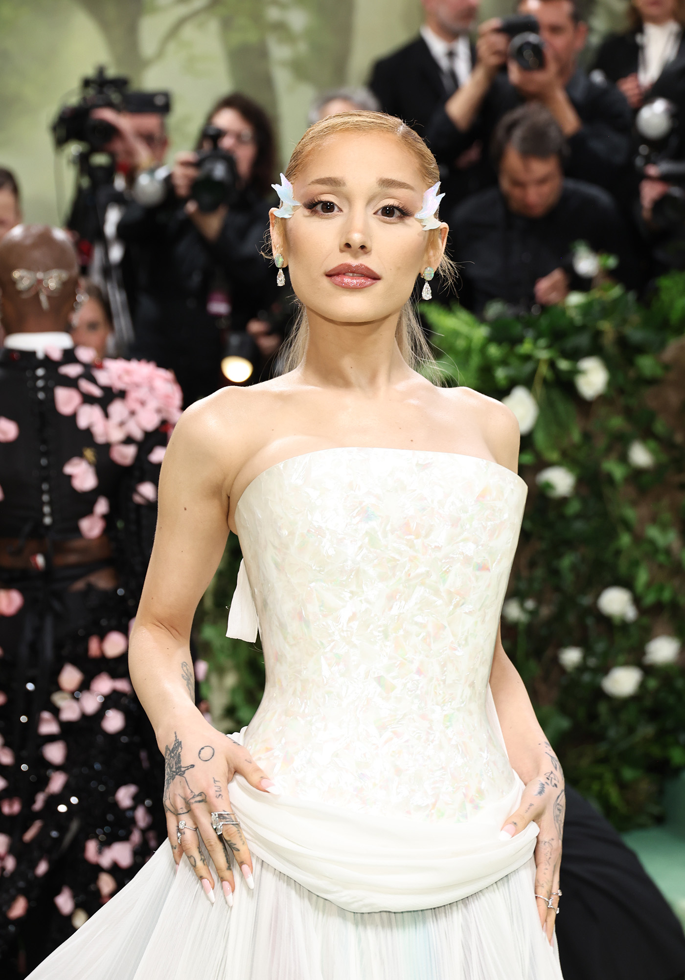 Ariana Grande w eleganckiej, perłowej sukni z kwiatowymi detalami, na tle zieleni i fotografów.