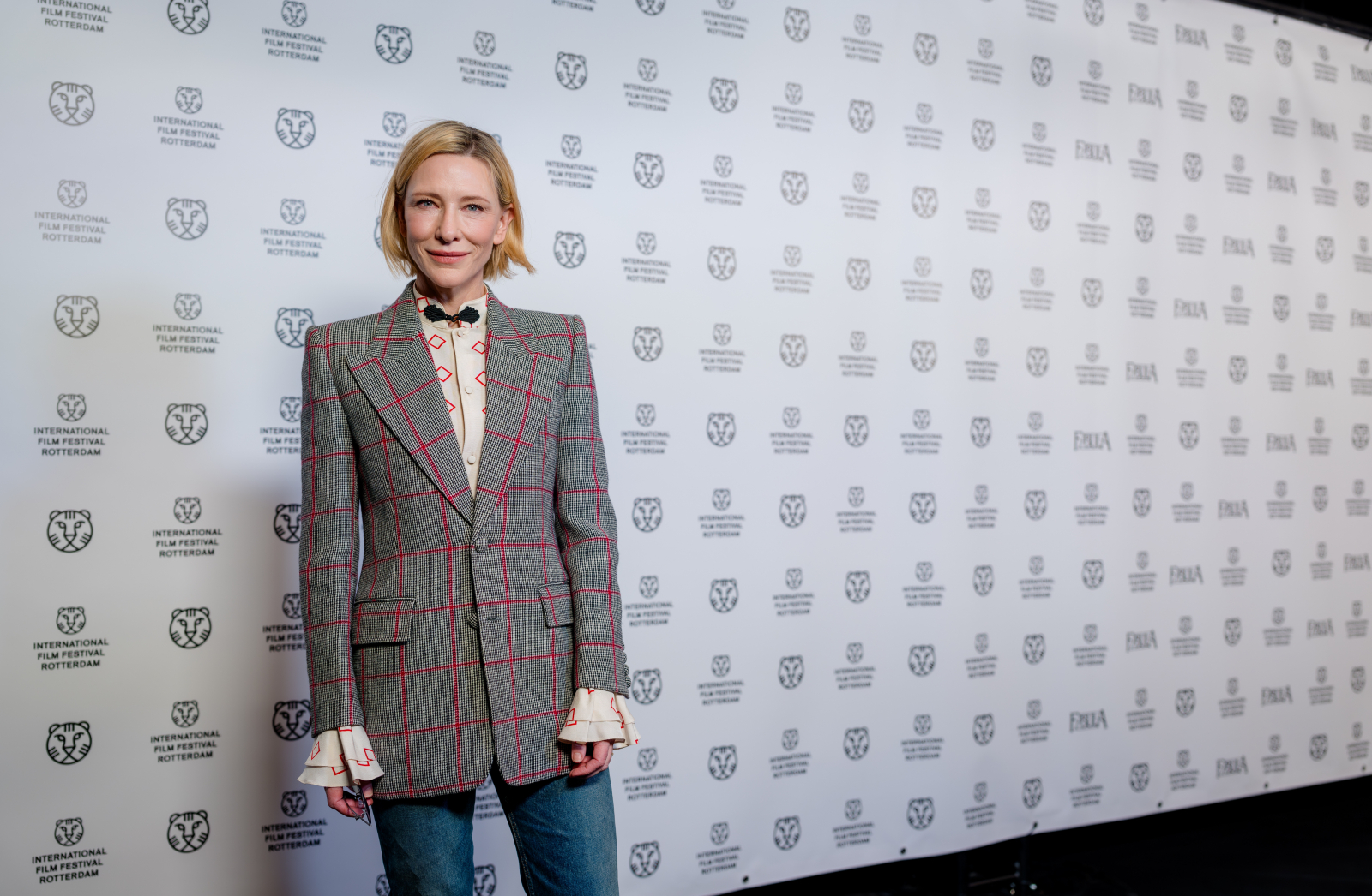 Cate Blanchett w eleganckim, szarym garniturze w kratę i dżinsach, pozująca na tle z logo festiwalu filmowego.