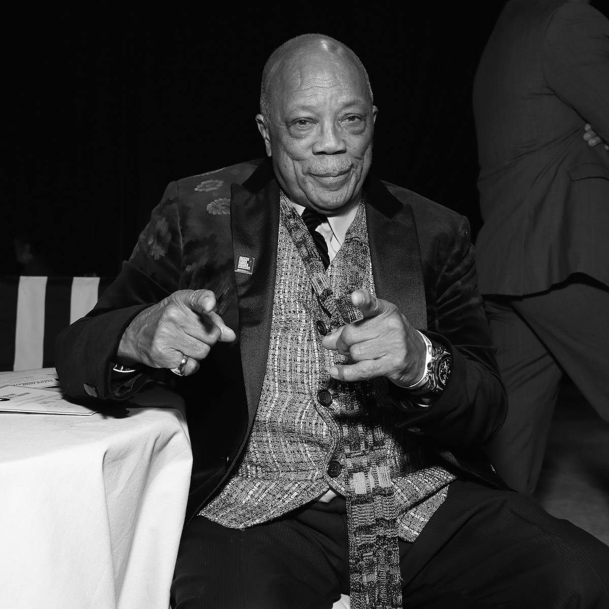 Quincy Jones, ubrany w elegancki garnitur, uśmiecha się i wskazuje w stronę obiektywu.