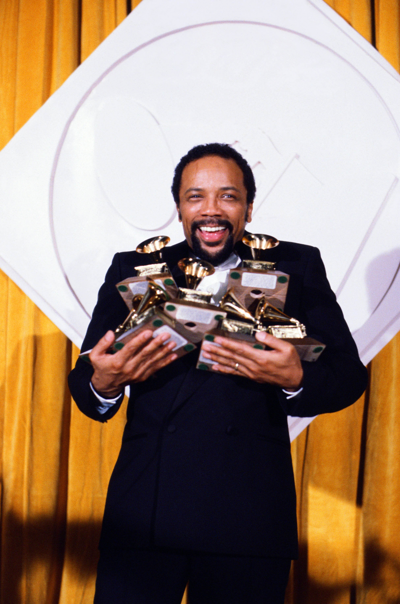 Quincy Jones trzyma w rękach wiele nagród Grammy, uśmiechając się na tle żółtej zasłony.