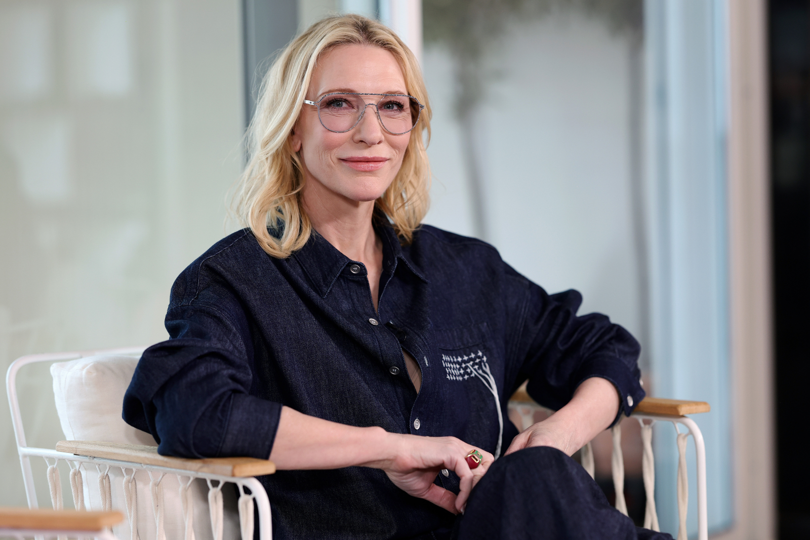 Cate Blanchett w okularach i granatowej koszuli, siedzi na białym fotelu. Uśmiecha się w kameralnej atmosferze.