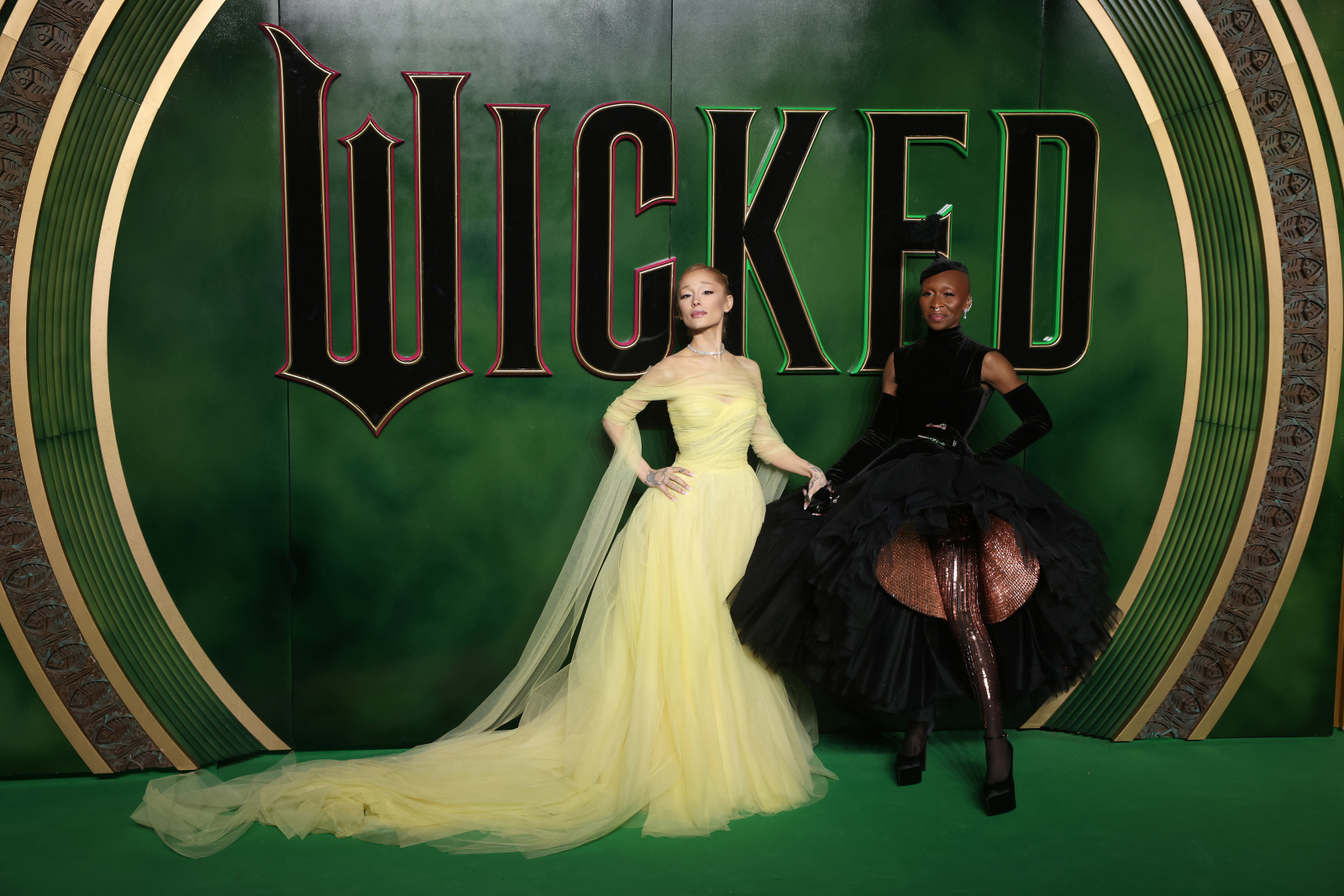 Ariana Grande i Cynthia Erivo pozujące na tle zielonego logo „Wicked&quot;, jedna w żółtej sukni, druga w czarnej, eleganckiej kreacji.