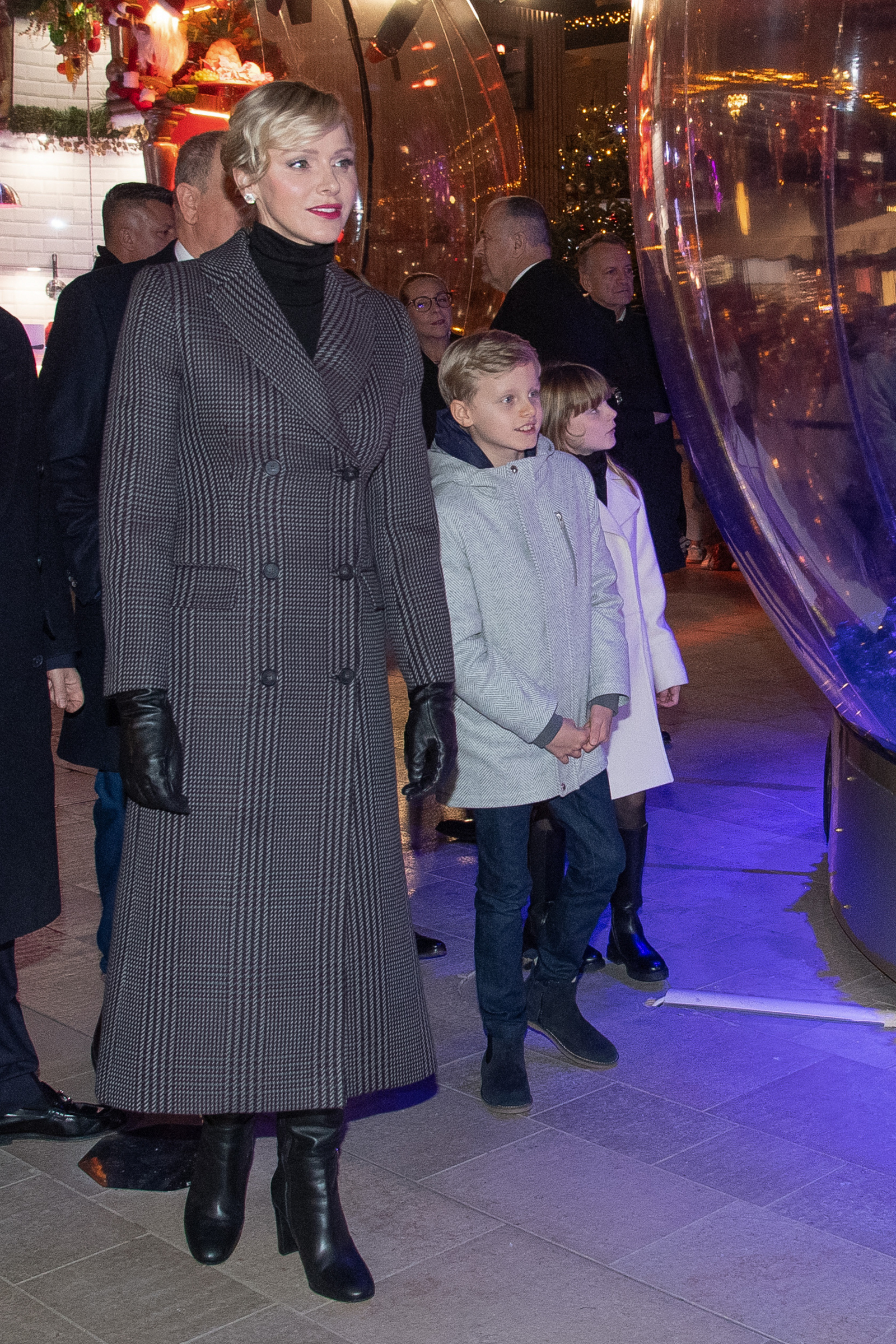 Księżna Charlene z Monako w eleganckim płaszczu i rękawiczkach, stoi obok dzieci, przy iluminacjach świątecznych.