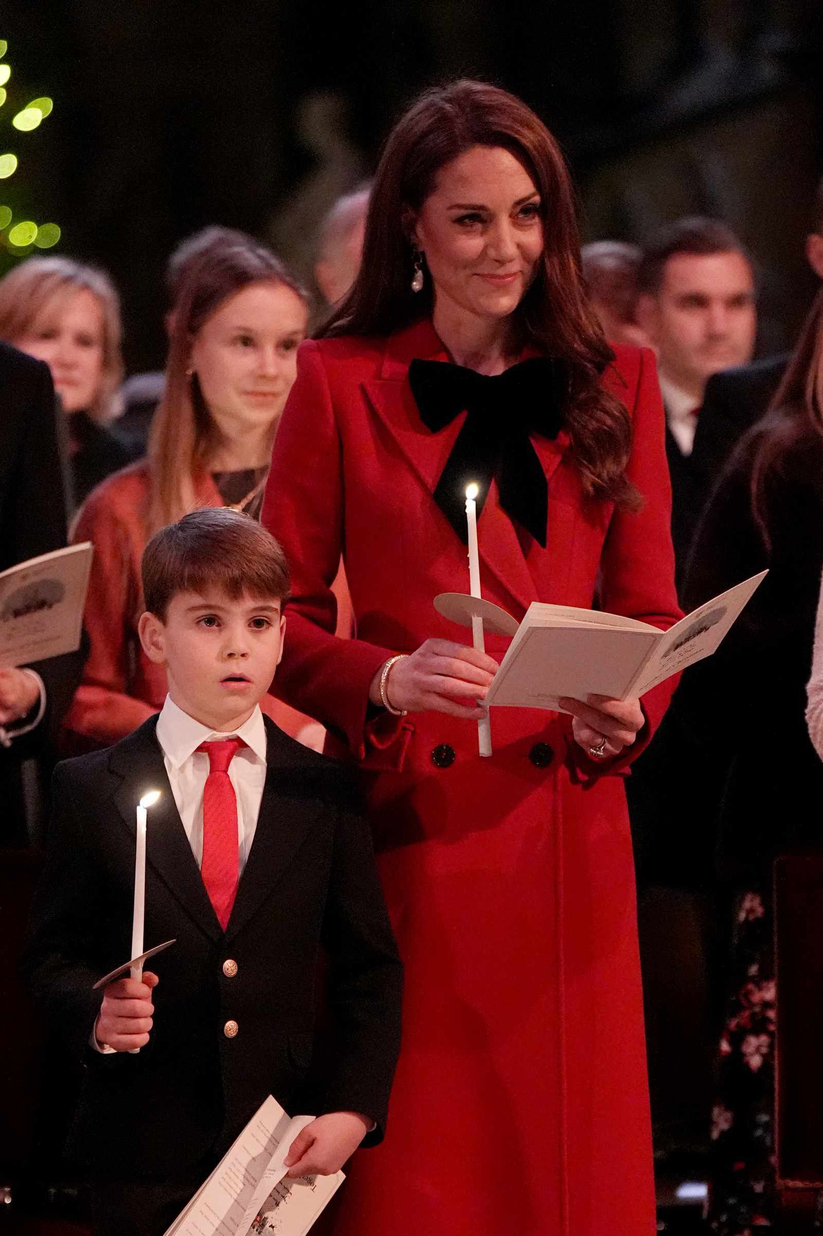 Księżna Kate w czerwonym płaszczu trzymająca świeczkę i książkę oraz książę George w garniturze z czerwoną krawatem, również ze świeczką.