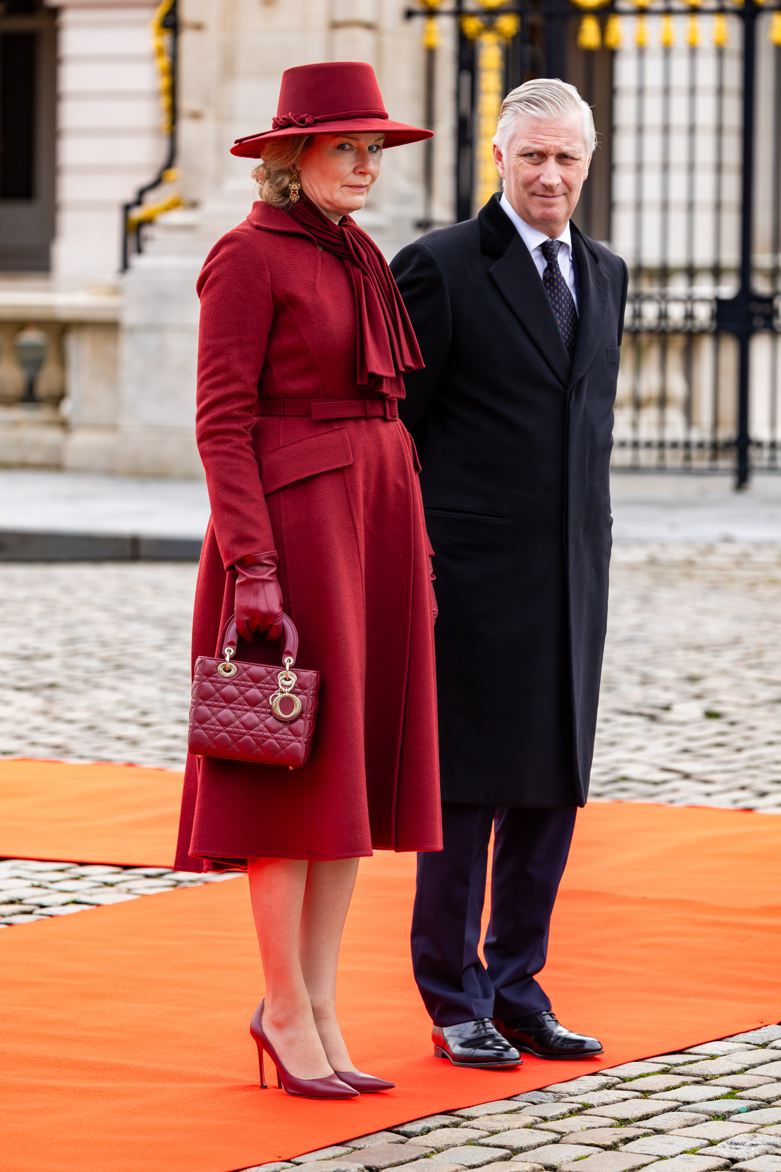 Królowa Matylda z Belgi w eleganckim, czerwonym płaszczu i kapeluszu stoi obok mężczyzny w czarnym płaszczu, na pomarańczowym dywanie.