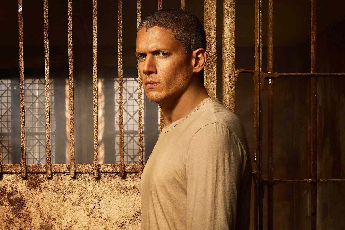 Wentworth Miller, wcielający się w rolę Michaela Scofielda,  stoi przy kratowej barierze, ma na sobie beżową koszulkę i patrzy poważnie w stronę kamery.