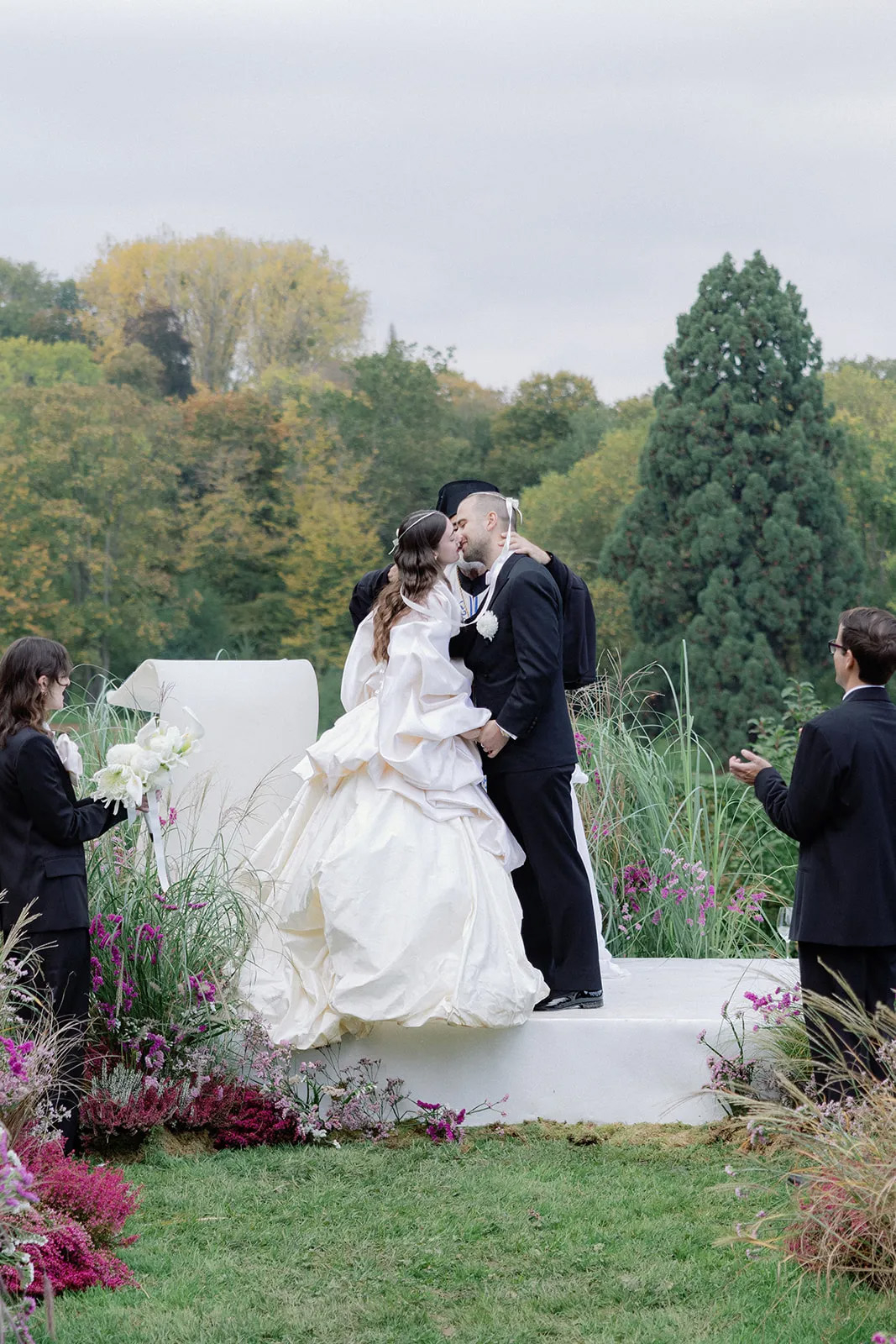 Para młoda w eleganckich strojach całuje się w plenerze, otoczeni zielenią oraz kwiatami.