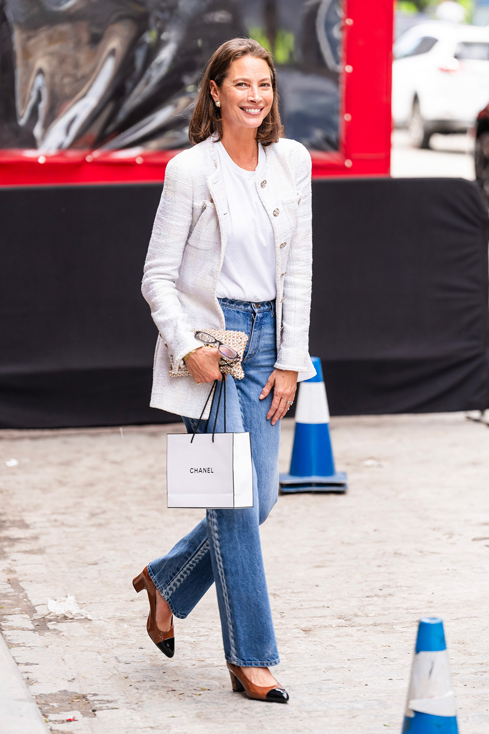 Christy Turlington w klasycznych jeansach, białej bluzce i eleganckiej marynarce, trzyma torebkę Chanel.