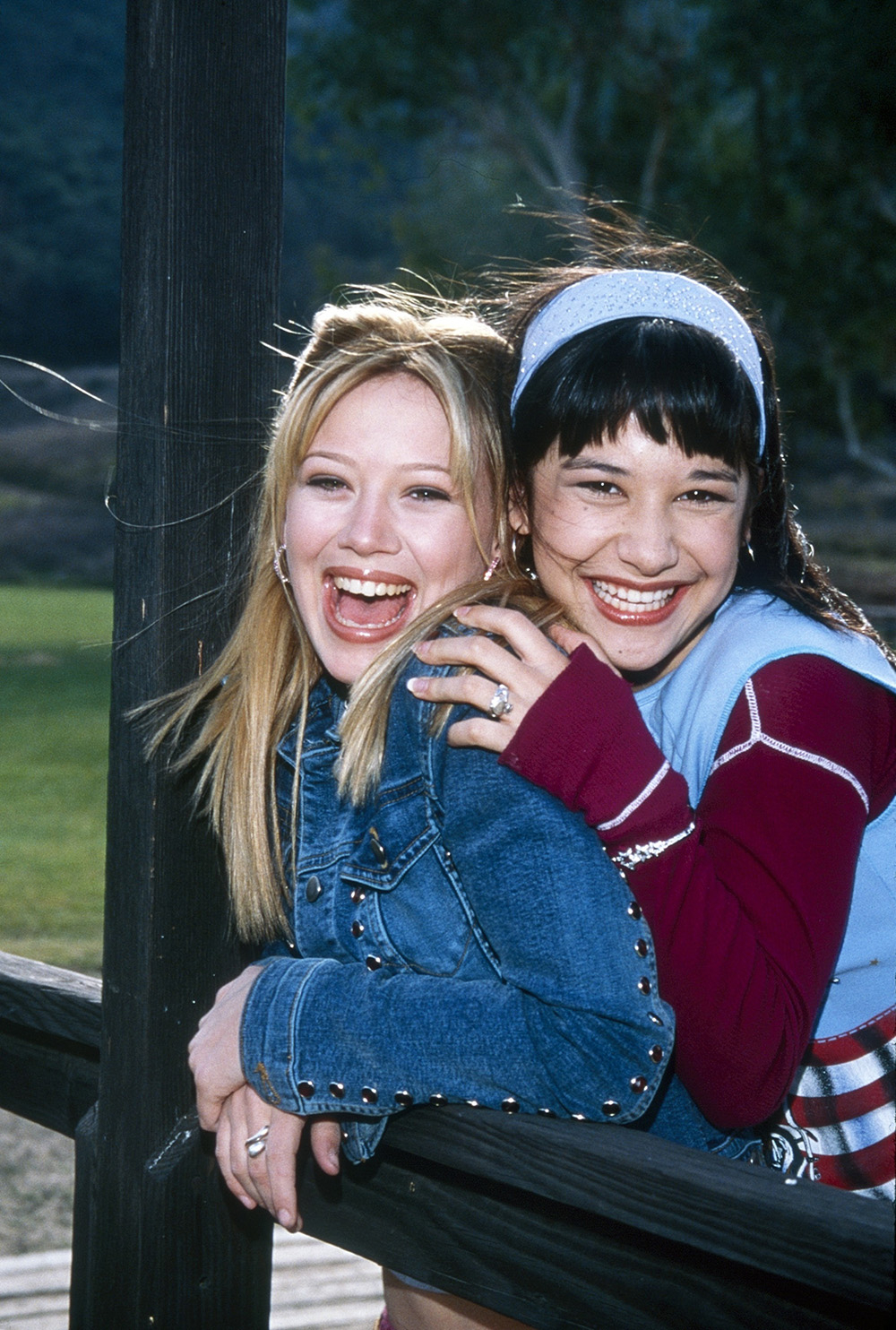 Lizzie McGuire i przyjaciółka uśmiechają się radośnie, trzymając się drewnianego słupa na tle zielonego krajobrazu.