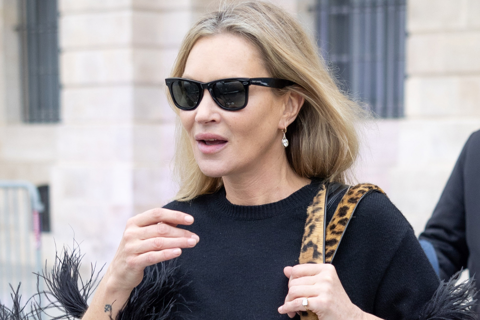 Kate Moss w czarnej bluzce z piórami, w okularach przeciwsłonecznych, ma siwe pasma we włosach.