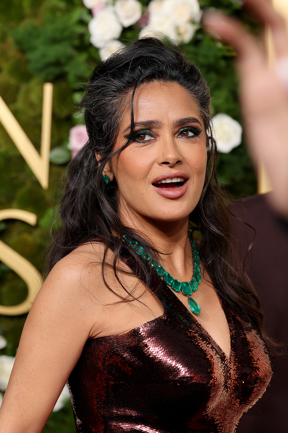 Salma Hayek w błyszczącej sukience z dekoltem, z naszyjnikiem z zielonych kamieni, na tle dekoracji kwiatowych.