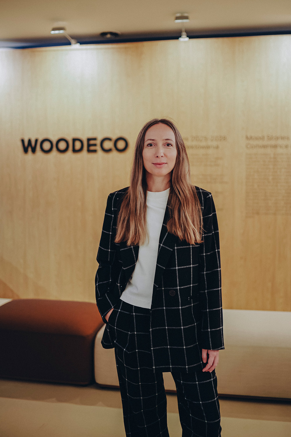 Maja Ganszyniec w czarnym garniturze w kratę stoi przed drewnianą ścianą z napisem "WOODDECO". W tle widoczna sofa.