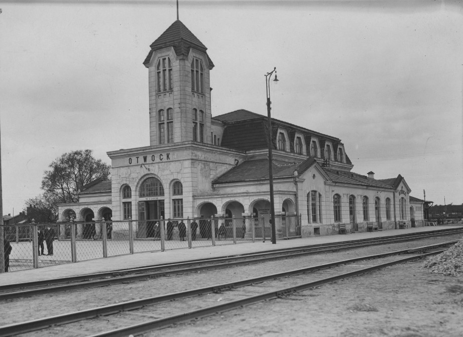 Dworzec kolejowy w Otwocku, z charakterystyczną wieżą i architekturą, otoczony pociągami i ludźmi.