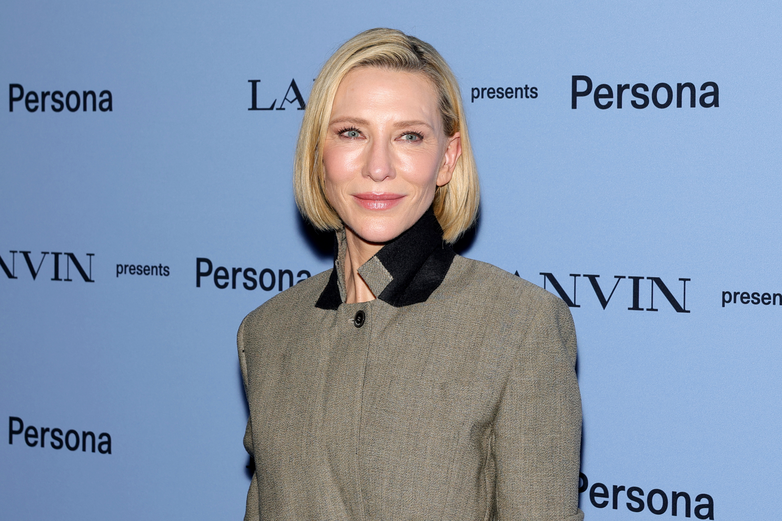 Cate Blanchett w eleganckim płaszczu, uśmiechając się na tle błękitnej ściany z napisami.