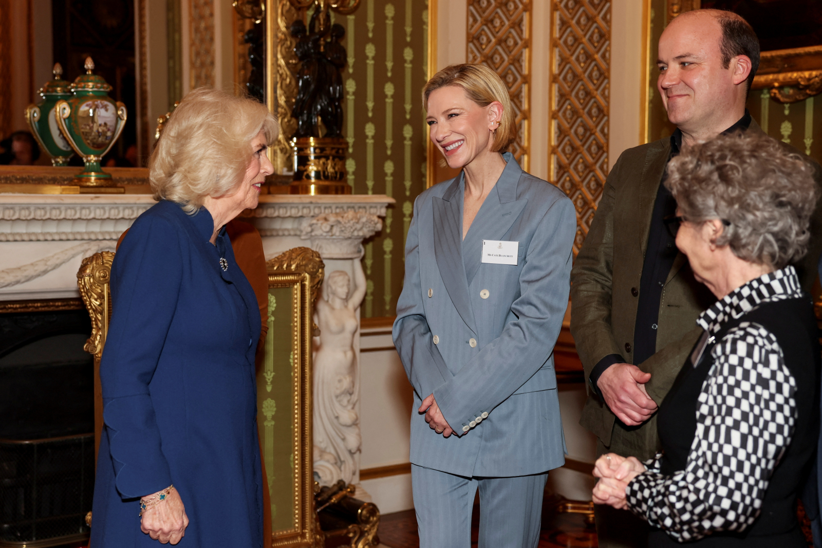 Królowa Kamila rozmawia z Cate Blanchett w eleganckim wnętrzu, obok stoją dwie inne osoby. Cate Blanchett ma na sobie garnitur w odcieniu gołębiej szarości.