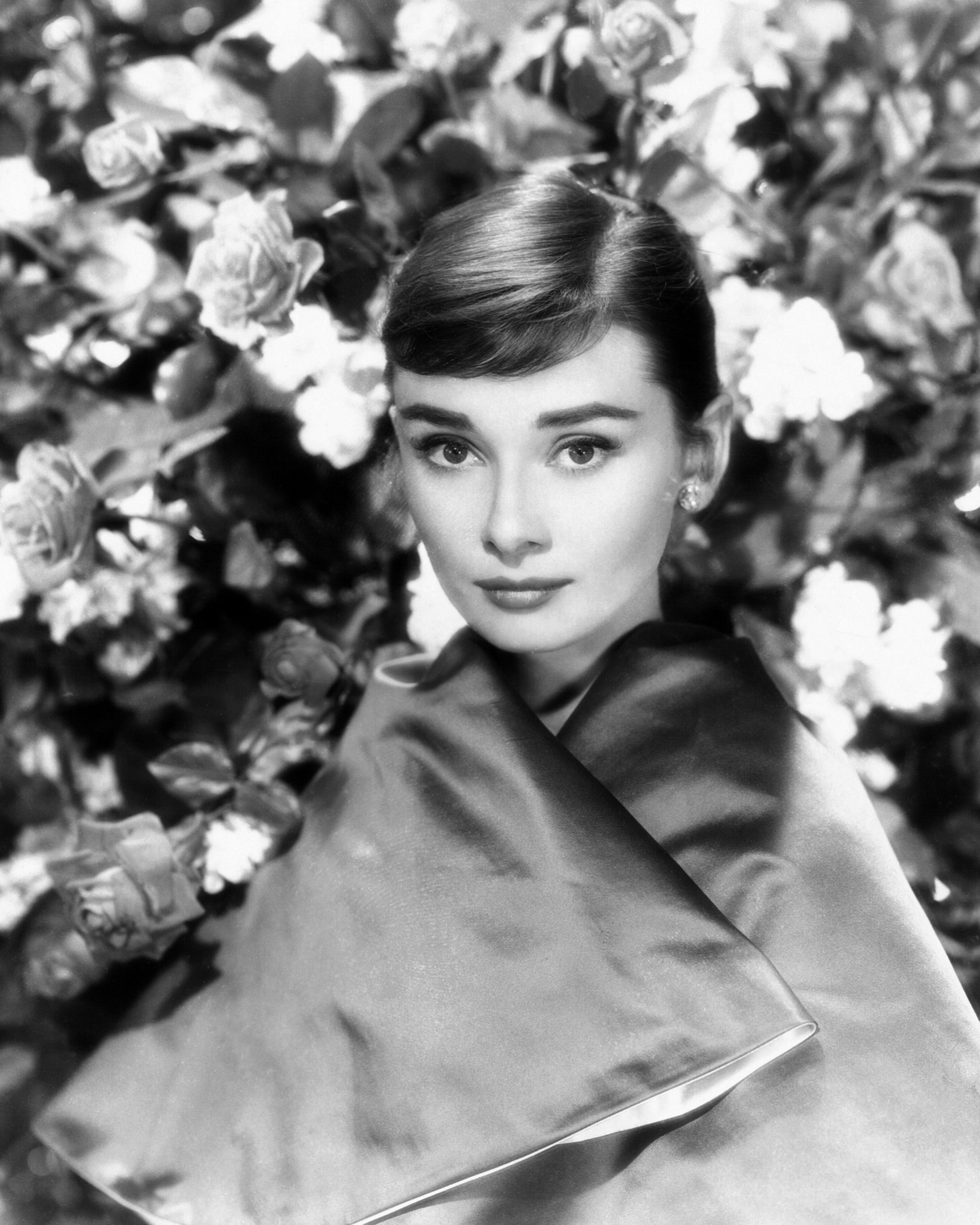 Audrey Hepburn w eleganckim stroju, otoczona kwiatami róż, z wyrazistym makijażem i delikatnym uśmiechem.