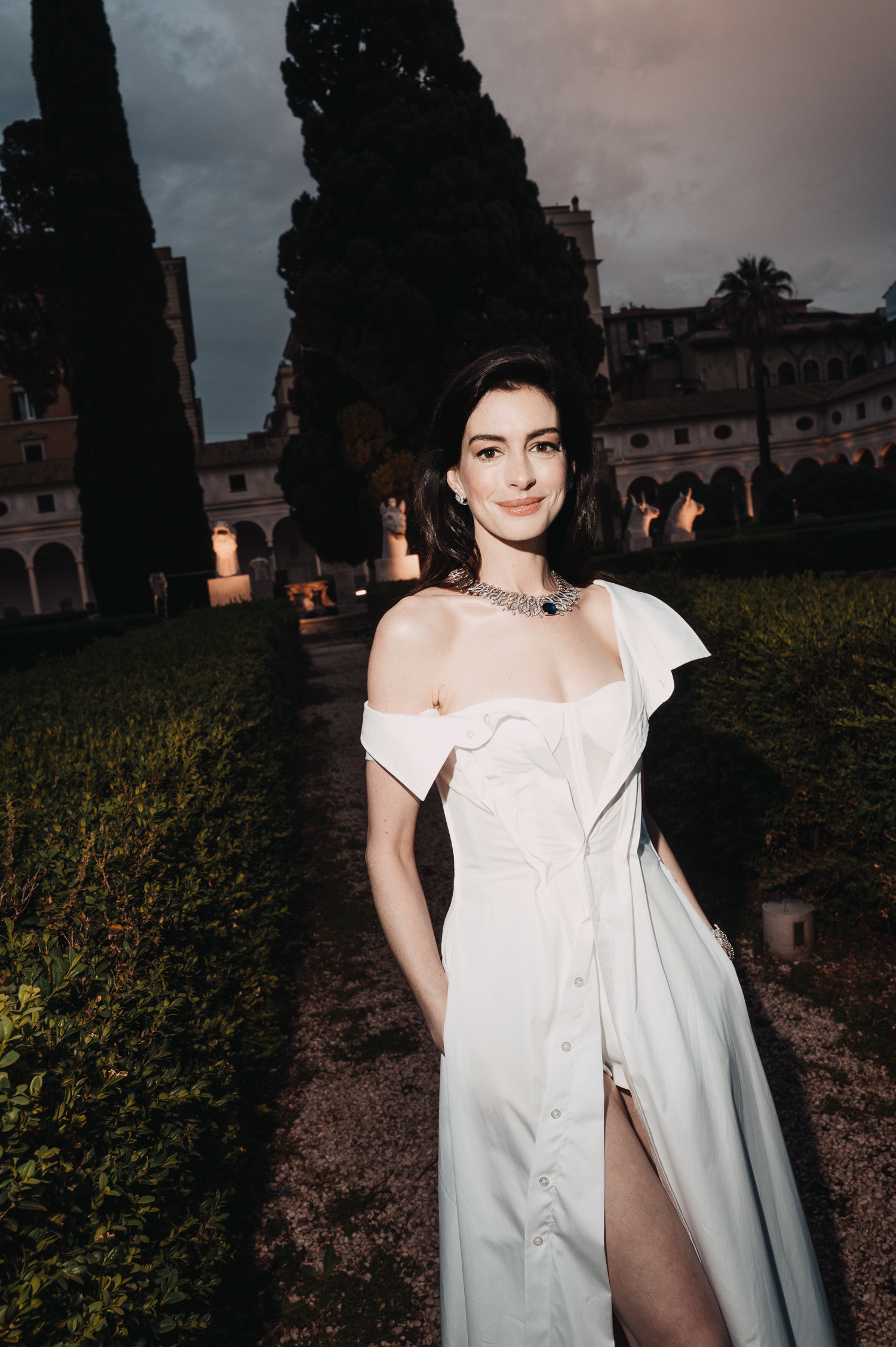 Anne Hathaway ubrana przez stylistkę Erin Walsh, w białej sukni, stoi w ogrodzie otoczonym drzewami.