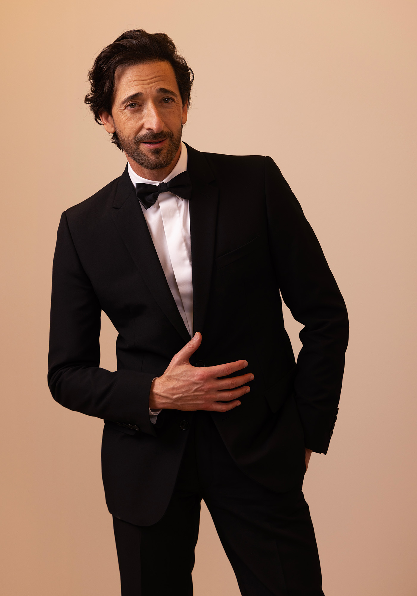 Adrien Brody w czarnym garniturze z muszką, stoi z jedną ręką na brzuchu, uśmiechając się. Tło w jasnym odcieniu.