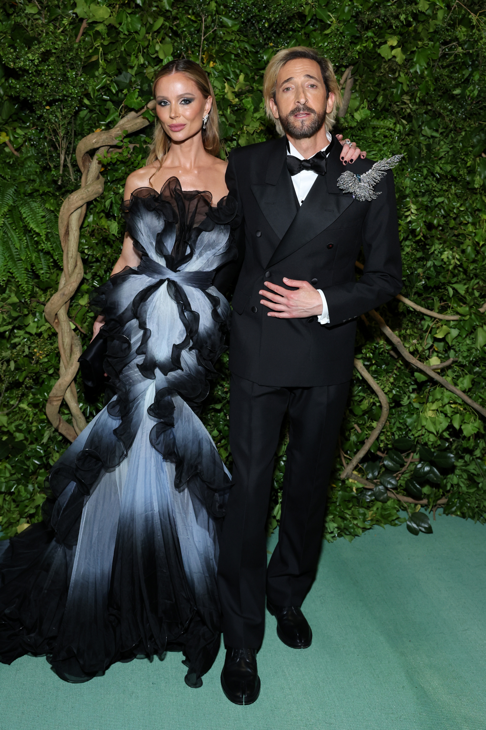 Adrien Brody i modelka Georgina Chapman pozują w eleganckich strojach, tło z roślinnością.