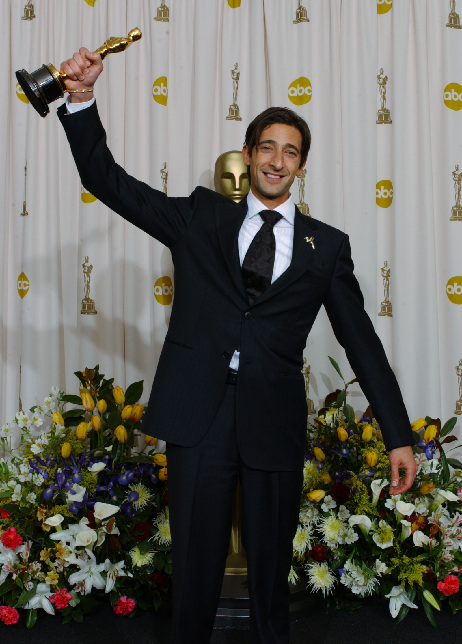 Adrien Brody trzyma Oscara, uśmiechając się szeroko, w otoczeniu kolorowych kwiatów.