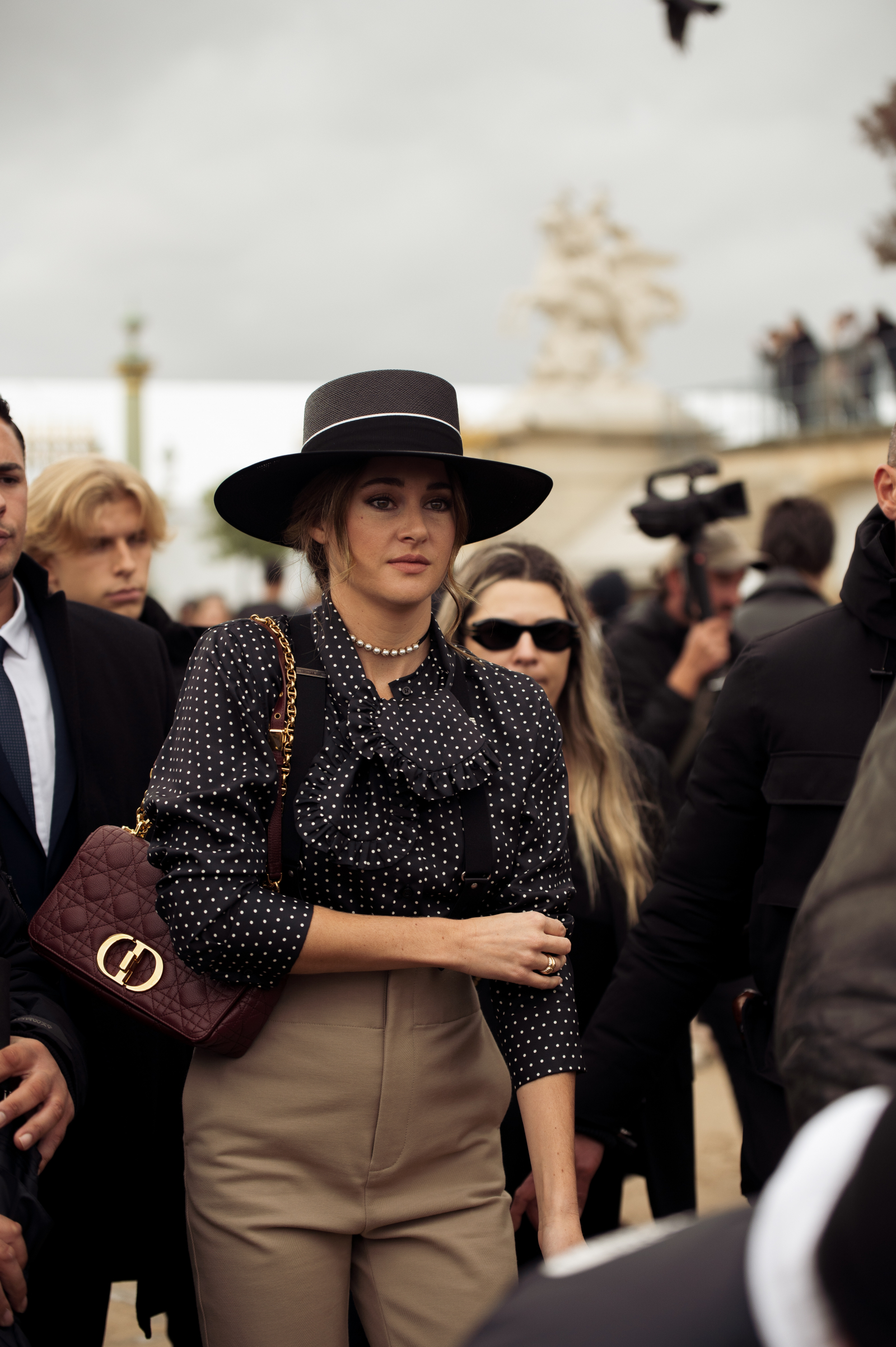 Aktorka Shailene Woodley w eleganckim stroju, bluzce w groszki, kapeluszu i z torebką, otoczona przez grupę ludzi na tle architektury.