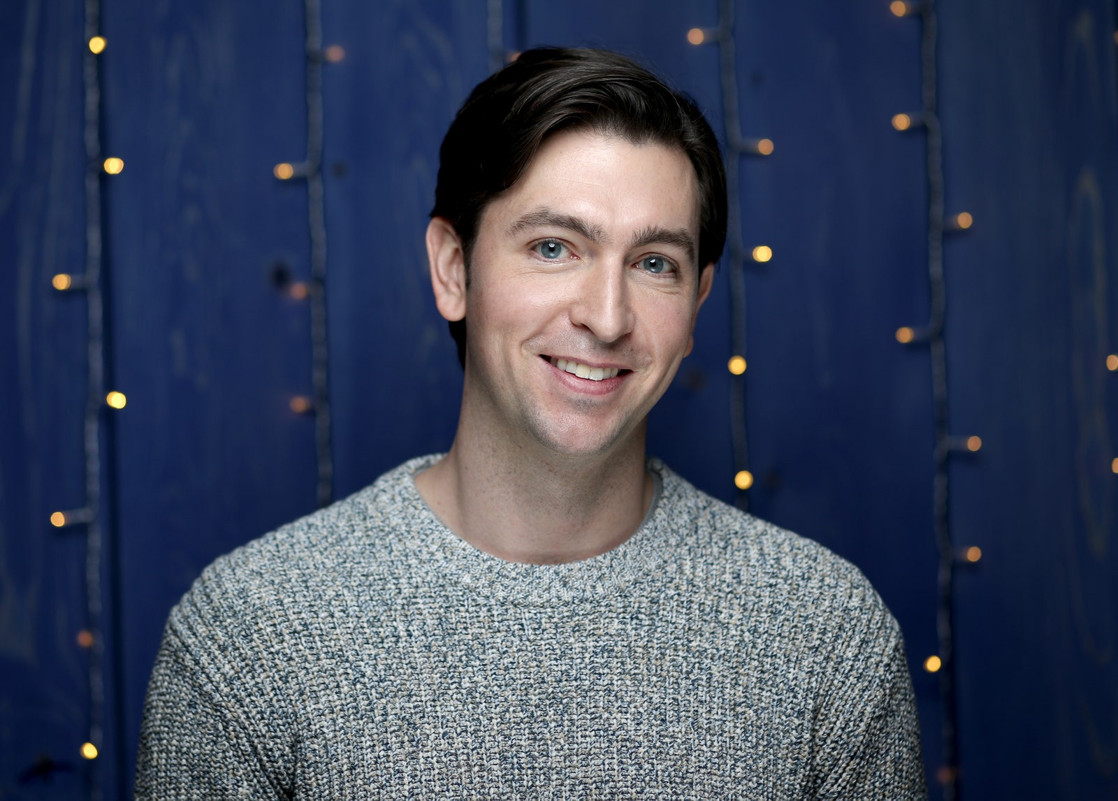 Nicholas Braun w szarym swetrze na niebieskim tle ze światełkami