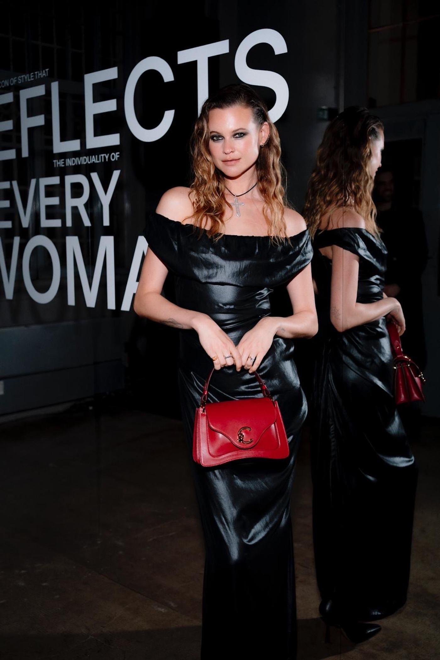 Behati Prinsloo w czarnej, błyszczącej sukni, z czerwoną torebką Coccinelle C-ME, stoi przed napisem "Reflects Every Woman".