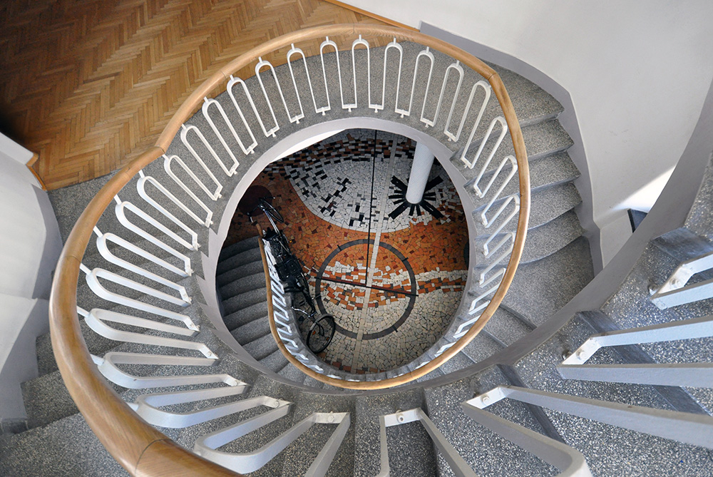 Widok na spiralne schody z drewnianym balustradą, na dole mozaika z rowerem na tle.