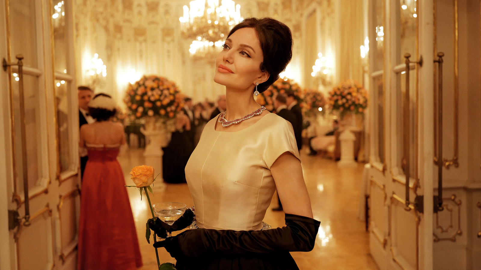 Angelina Jolie w eleganckiej sukni na przyjęciu, trzymająca różę, z filmu „Maria”. W tle luksusowa sala.