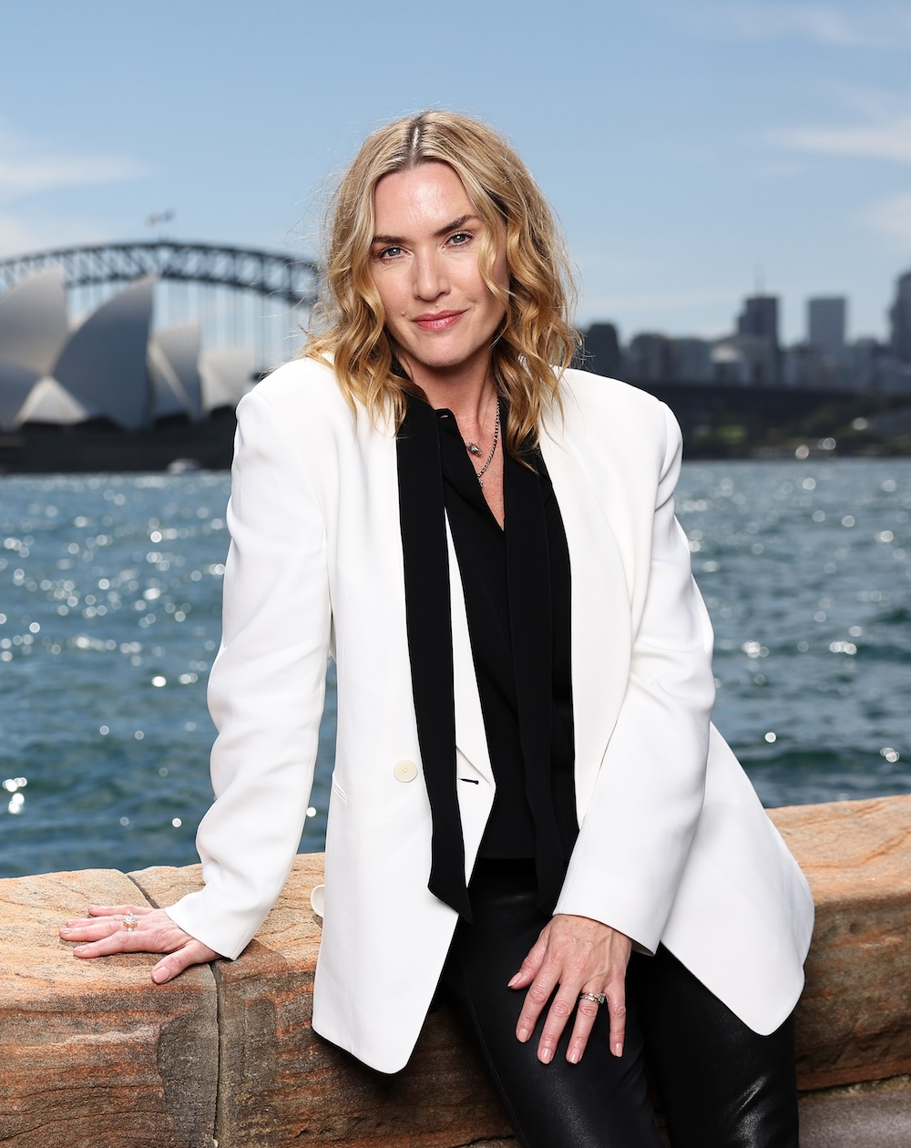 Kate Winslet w białej marynarce damskiej, nad wodą, z Sydney w tle.