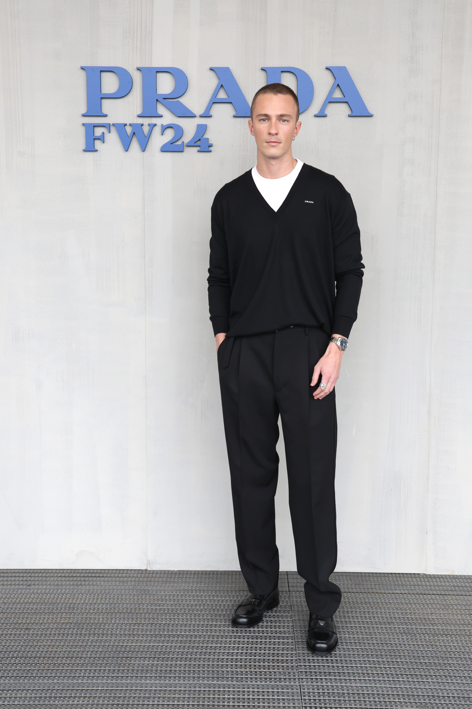 Drew Starkey w czarnej stylizacji, stoi przed napisem "PRADA FW24" na szarej ścianie.