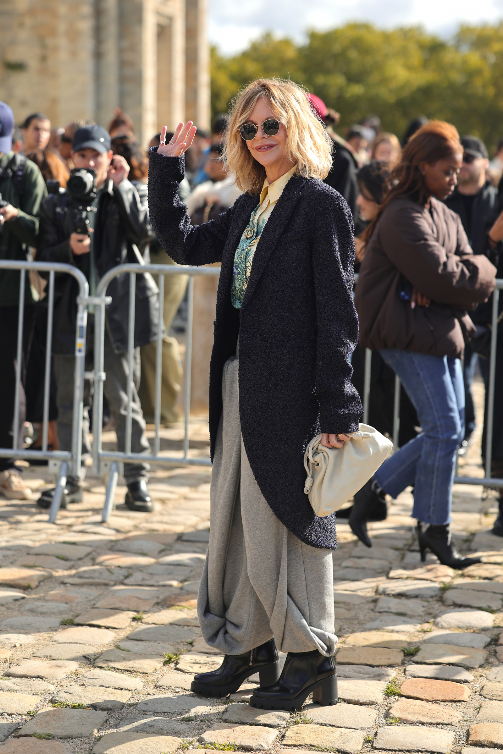 Aktorka Meg Ryan w szerokich spodniach i botkach na słupku, machająca do tłumu fanów.