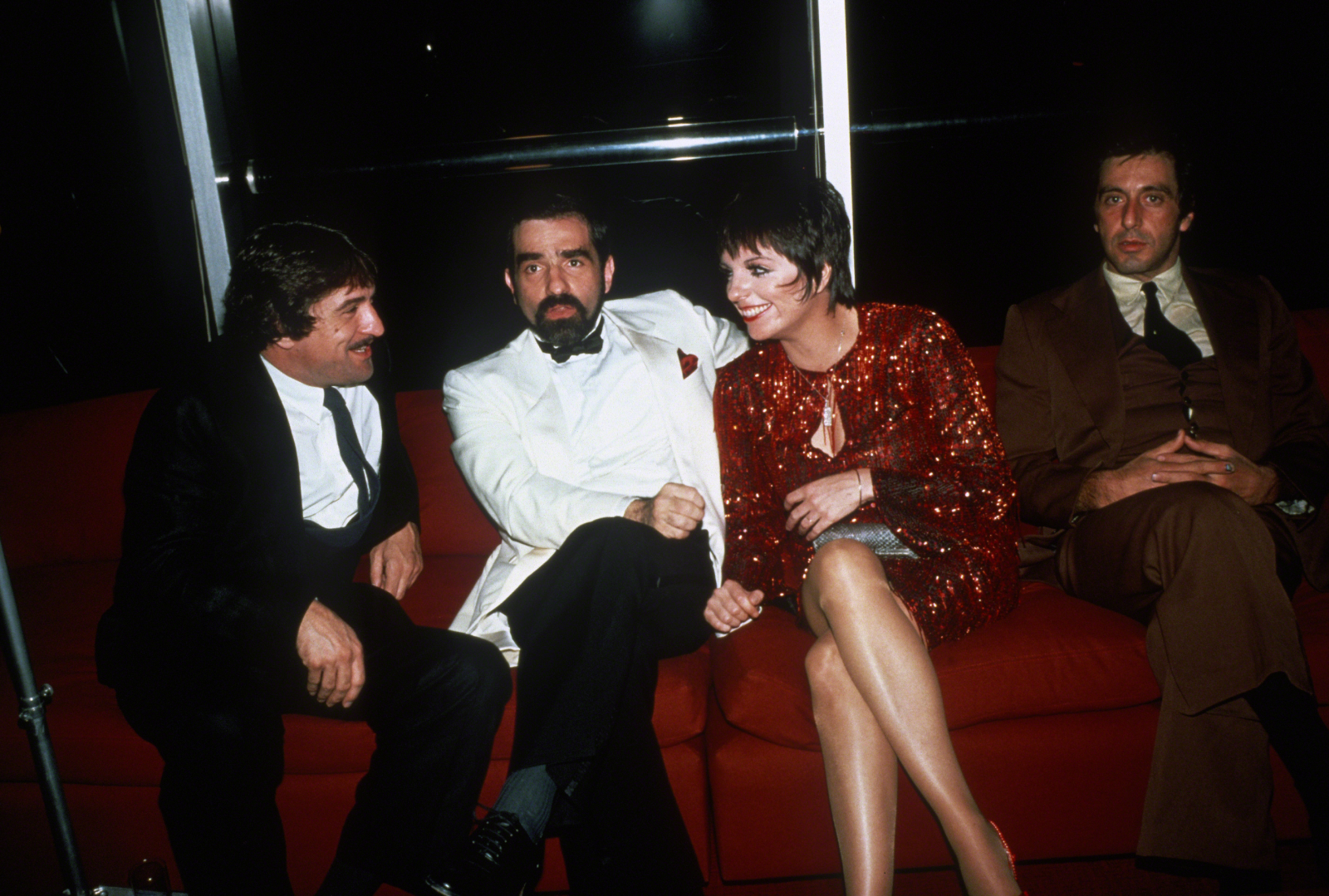 Robert De Niro i Al Pacino rozmawiają z Martinem Scorsese oraz Liza Minnelli na kanapie w eleganckim otoczeniu.