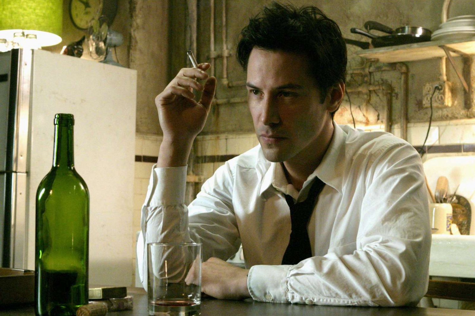 Keanu Reeves w scenie z „Constantine”, filmie z 2005 roku, siedzi przy stole z butelką i szklanką.