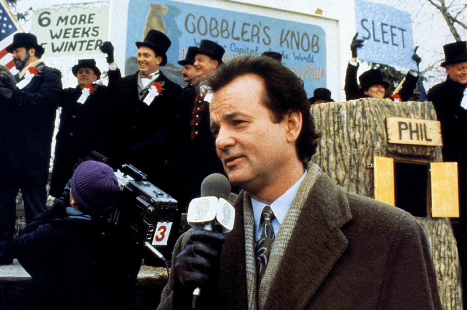 Bill Murray w roli reportera obok grupy mężczyzn w cylindrach, na tle wydarzenia z świetnego filmu „Dzień świstaka”.