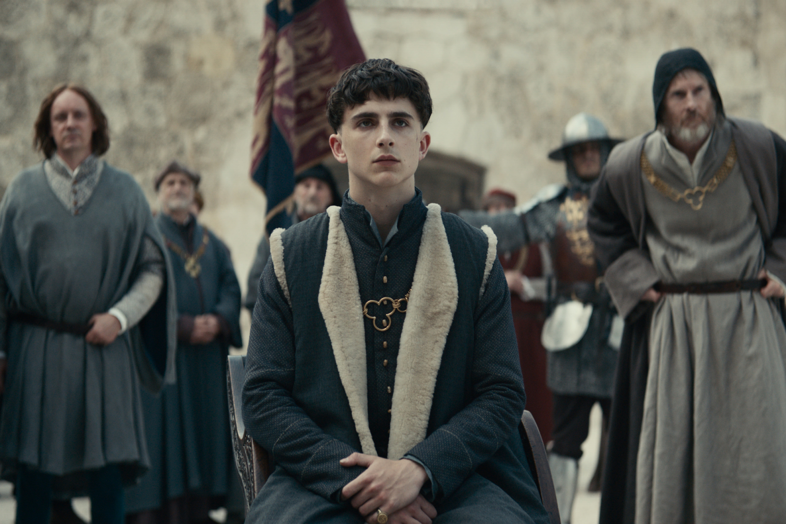 Timothée Chalamet w filmie Netflix „Król”, siedzący na tronie w otoczeniu doradców i wojskowych.