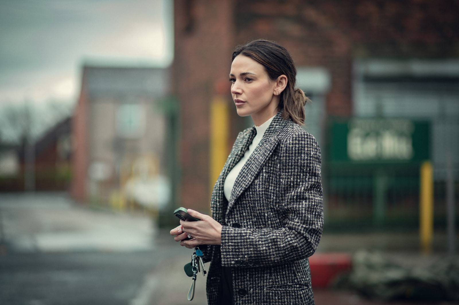 Kobieta w eleganckim płaszczu stoi na zewnątrz, trzymając klucze, w tle sceny z serialu Netflix „Już mnie nie oszukasz”.