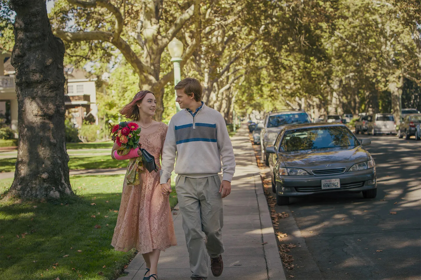 Kadr z filmu „Lady Bird”, reżyseria Greta Gerwig. Młoda para grana przez Saoirse Ronan i Lucasa Hedgesa spaceruje po ulicy, trzymając się za ręce i uśmiechając, w otoczeniu drzew.