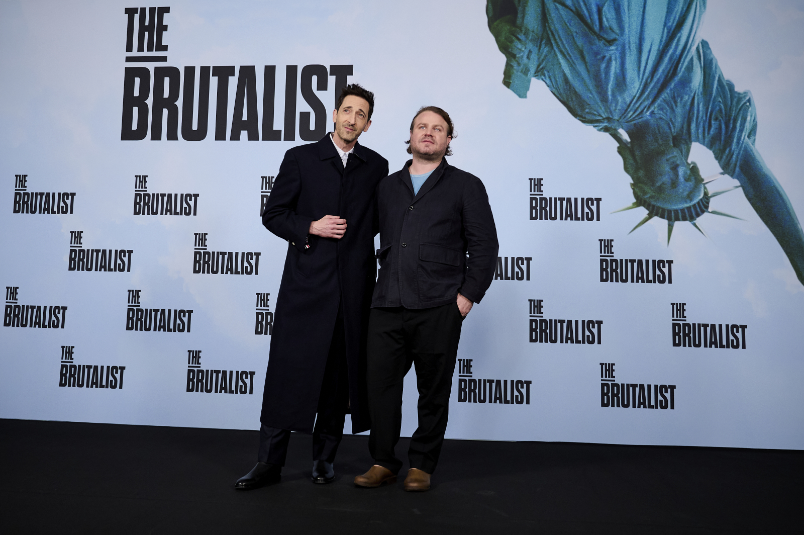 Adrien Brody i Brady Corbet na premierze filmu &quot;The Brutalist&quot;, w tle Statua Wolności oraz logo filmu.