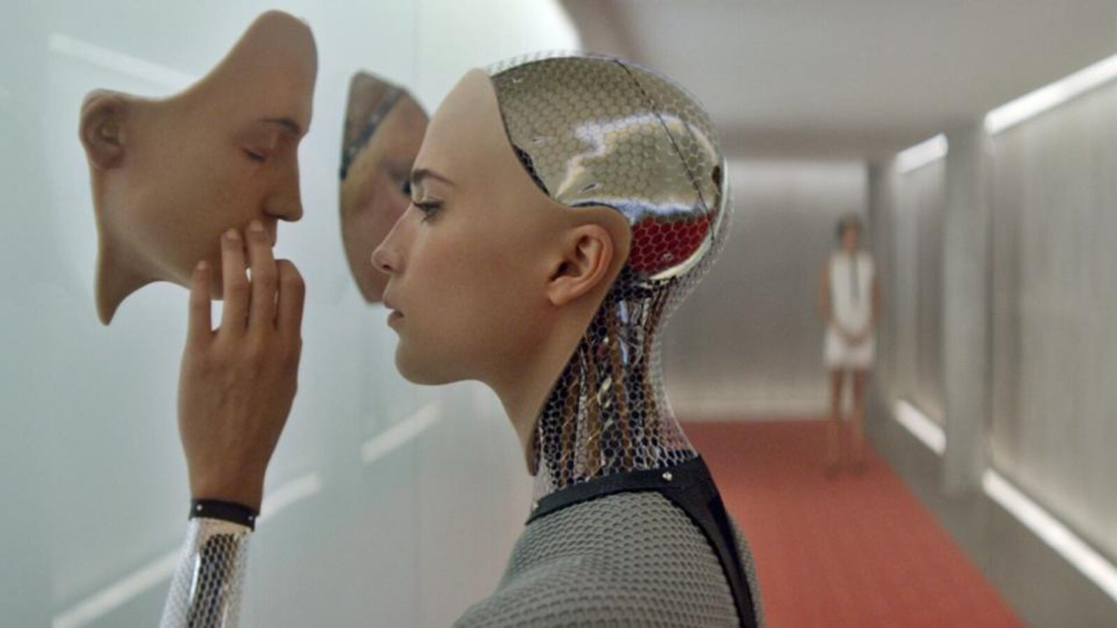 Kadr z filmu „Ex Machina” z 2014 roku w reżyserii Alexa Garlanda przedstawia kobiecego robota Avę (graną przez Alicię Vikander) dotykającego przezroczystej ściany, obok stoi druga postać.