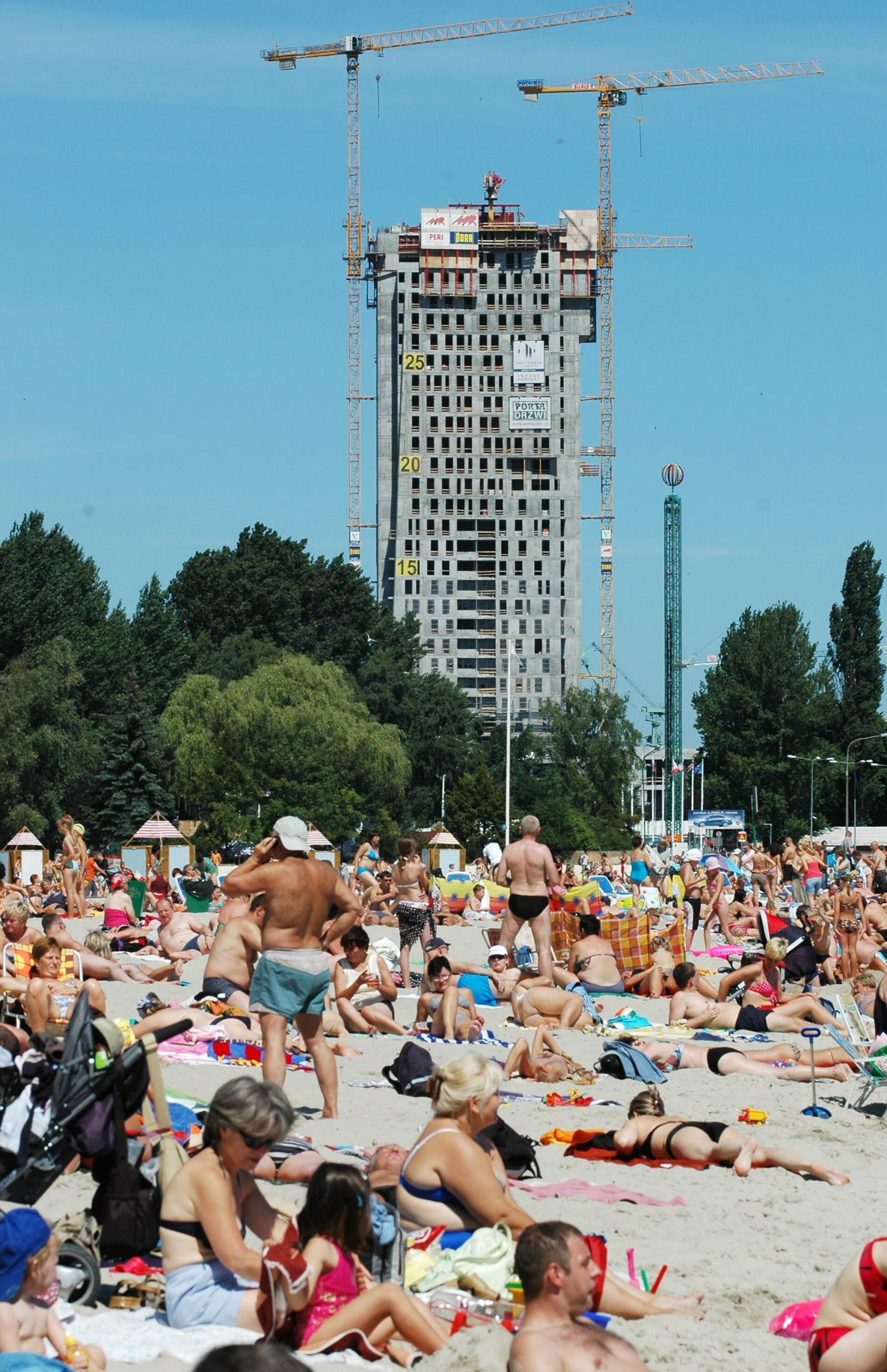 Plaża w Gdyni w słoneczny dzień, tłum ludzi opala się, w tle widać budowę wysokiego wieżowca.