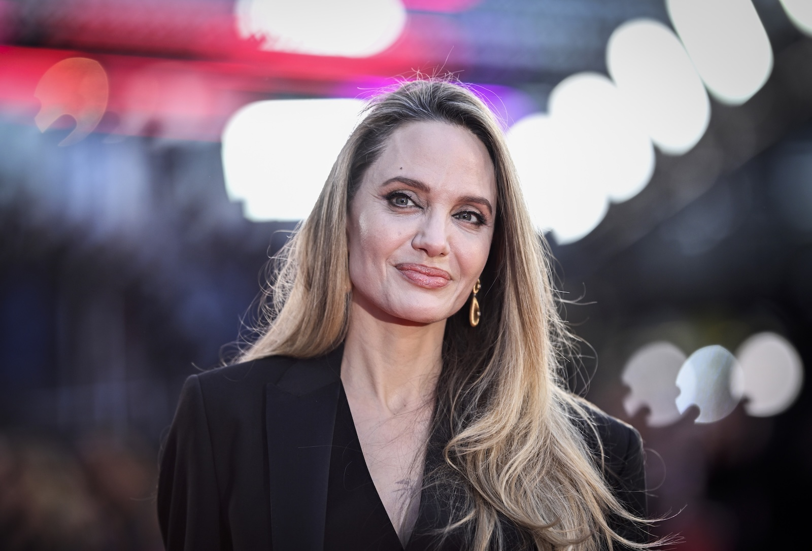 Angelina Jolie w eleganckim czarnym stroju, uśmiechając się na tle rozmytych świateł podczas premiery filmu "Maria" w Londynie.