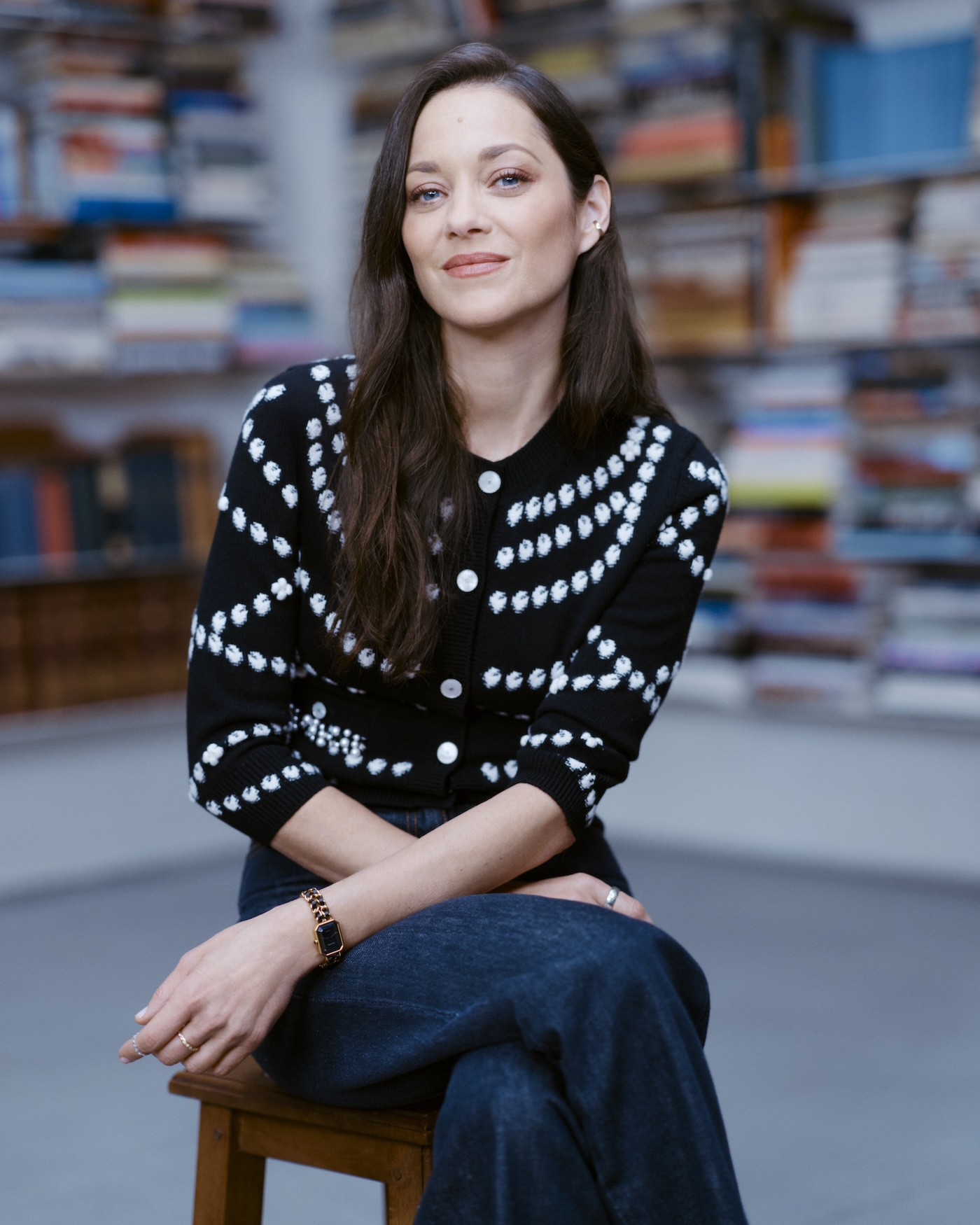 Marion Cotillard wczarnym kardiganie z białymi ozdobnikami i w błękitnych spodniach pozuje do zdjęcia siedząc na drewnianym stołku na tle biblioteczki.