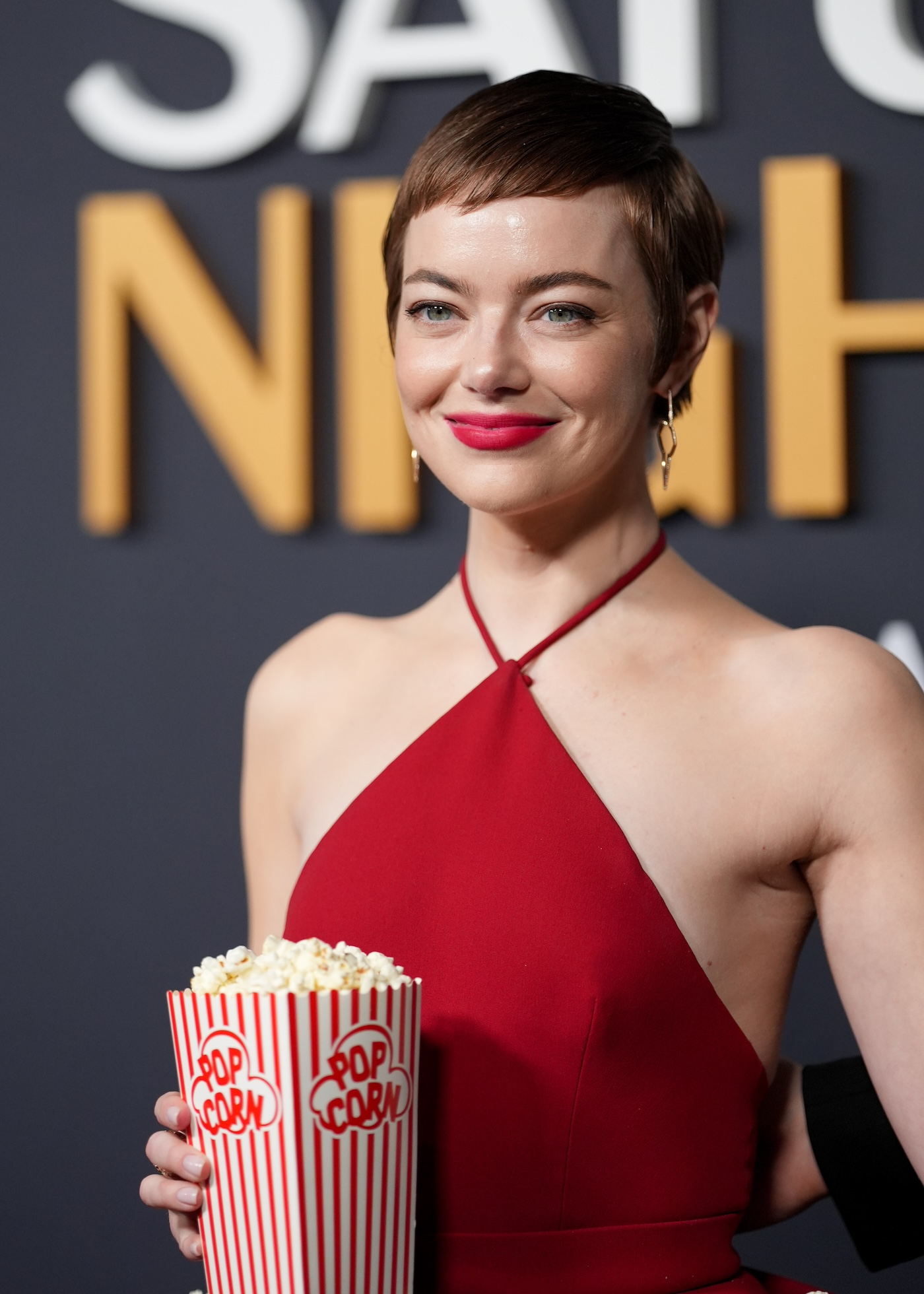 Emma Stone w czerwonej sukience z wiązaniem na szyi, trzyma w ręku papierowe pudełko z popcornem, ma krótkie włosy i wyrazisty makijaż z ustami podkreślonymi czerwoną szminką.