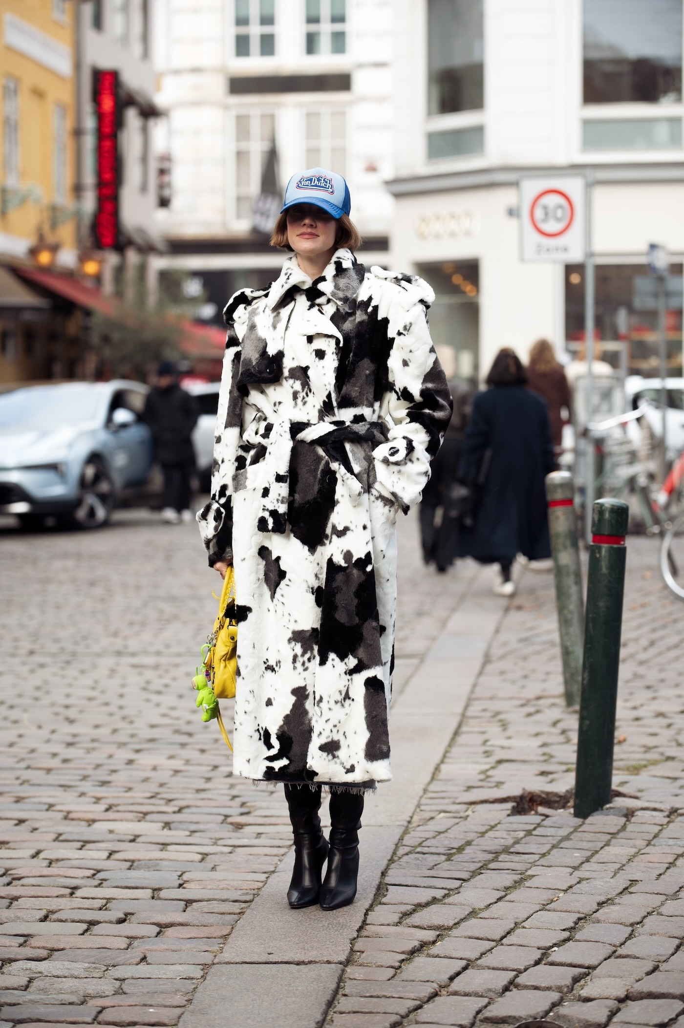 Marianne Theodorsen ma na sobie płaszcz w krowie łaty, intensywnie żółtą torbę z dodatkami, błękitną czapkę z daszkiem i czarne skórzane kozaki podczas Copenhagen Fashion Week.