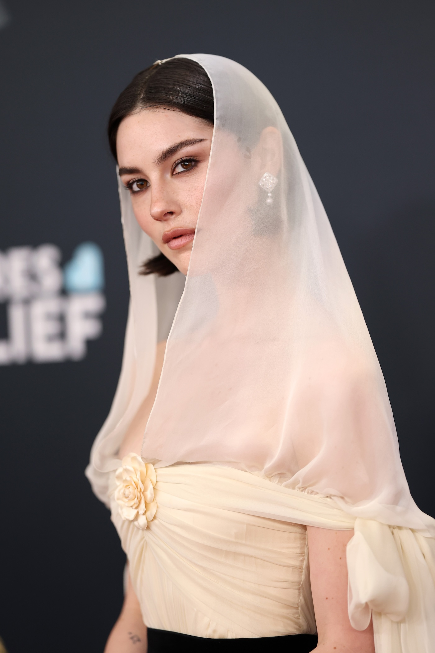 Gracie Abrams podczas rozdania nagród Grammy w 2025 r., w beżowej, szyfonowej sukni od Chanel z odkrytymi ramionami, ozdobną kamelię i eleganckimi wiązaniami na rękach, delikatnym welonem oraz czarnym paskiem.