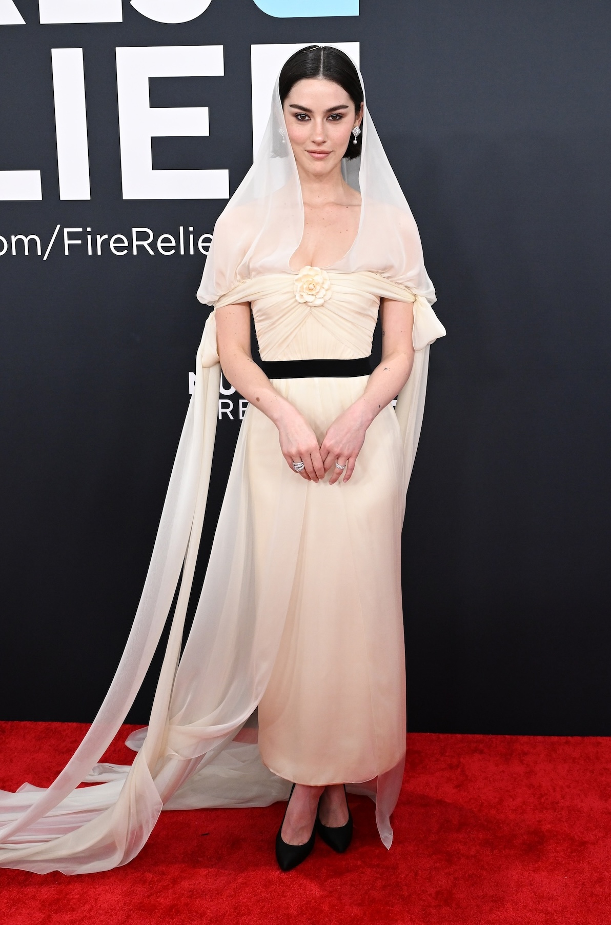 Gracie Abrams podczas rozdania nagród Grammy w 2025 r., w beżowej, szyfonowej sukni od Chanel z odkrytymi ramionami, ozdobną kamelię i eleganckimi wiązaniami na rękach, delikatnym welonem, czarnym paskiem i czarnymi szpilkami.