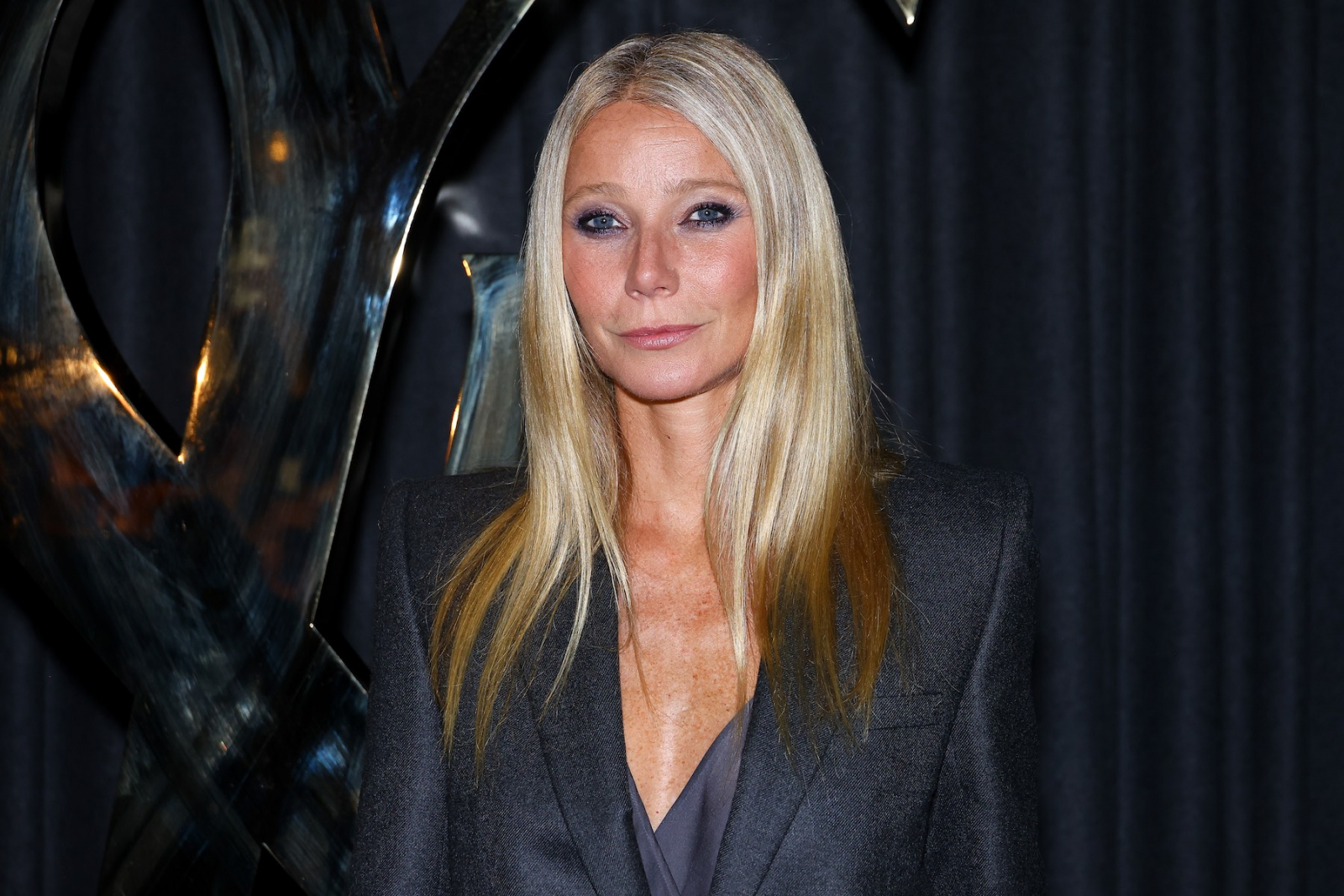 Gwyneth Paltrow w eleganckim garniturze, z długimi, prostymi włosami, stoi na tle ciemnej zasłony i metalowej rzeźby.