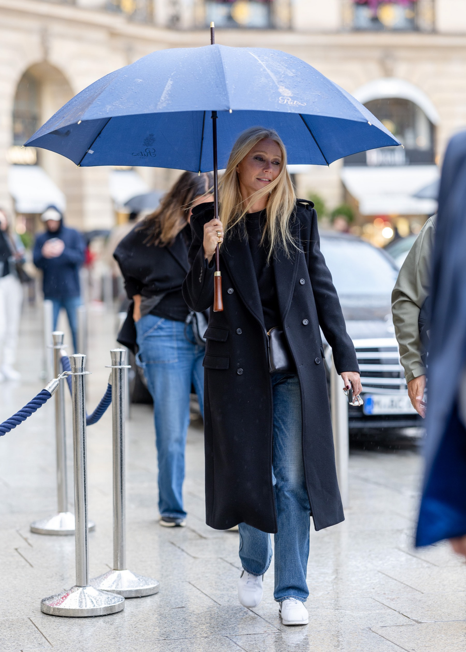 Gwyneth Paltrow w ciemnym płaszczu, dżinsach i białych butach, trzyma niebieski parasol, spacerując po deszczowej paryskiej ulicy.