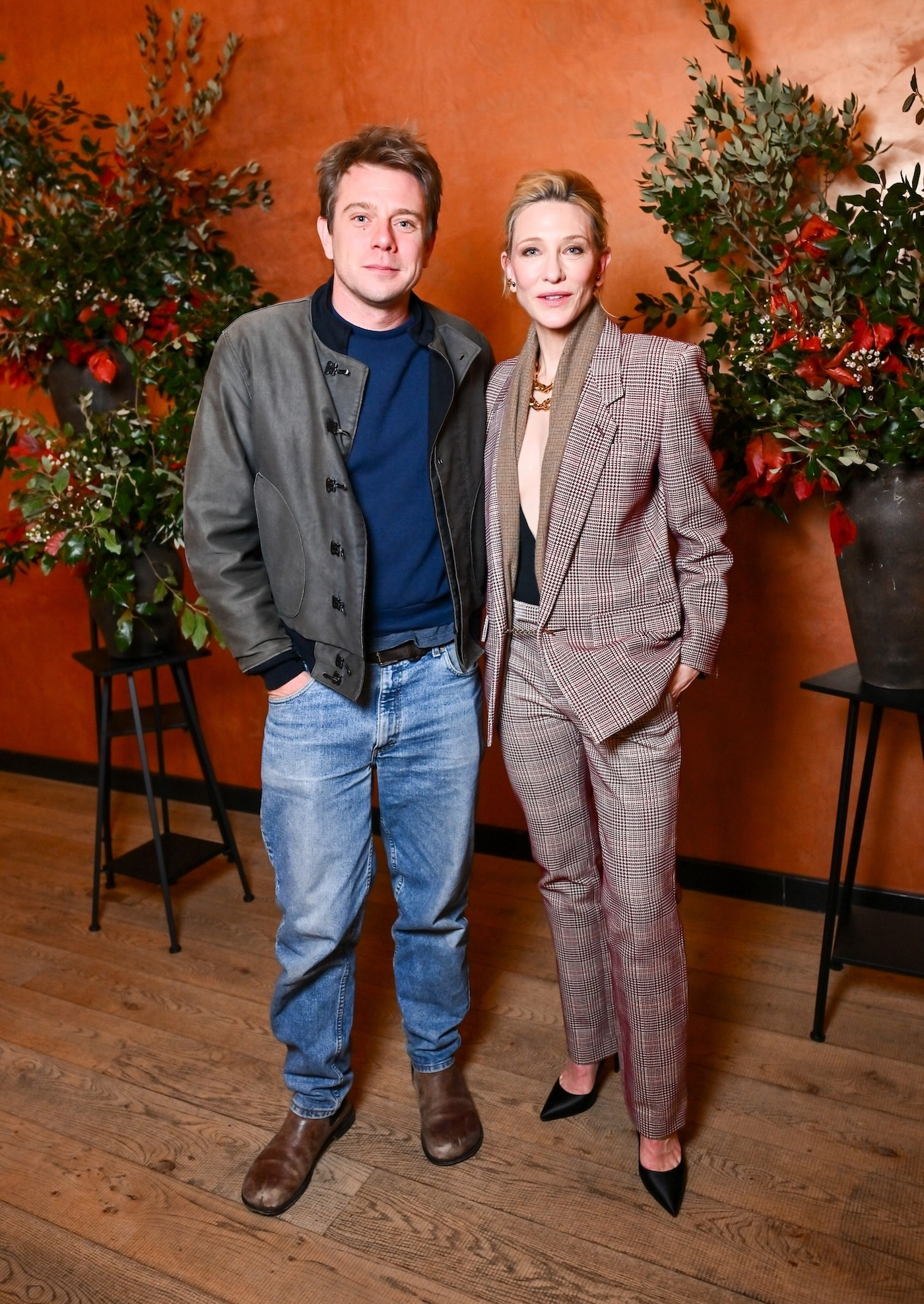 Jonathan Anderson i Cate Blanchett pozują wspólnie do zdjęcia podczas wydarzenia Vogue: Inventing the Runway" na tle rudej ściany i roślinnych dekoracji.